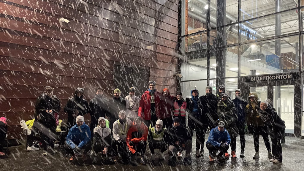 24 godt kledde løpere står sammen i et gruppebile utenfor et stort bygg i ganske tett snøvær.