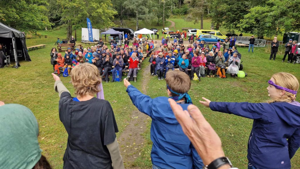 I forgrunnen står barn og viser til teknikertelt til venstre, mens publikum klapper.