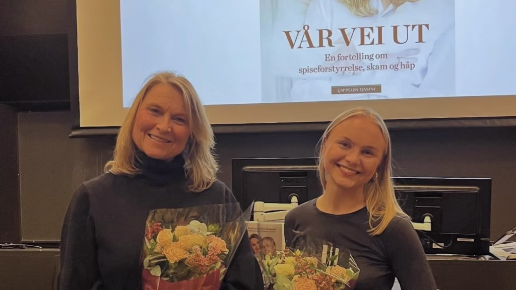 To kvinner, mor og datter, som har fått hver sin blomsterbukett. De står foran et lerret med bilde av boka "Vår vei ut". På forsiden av boka er det bilde av de samme to kvinnene.