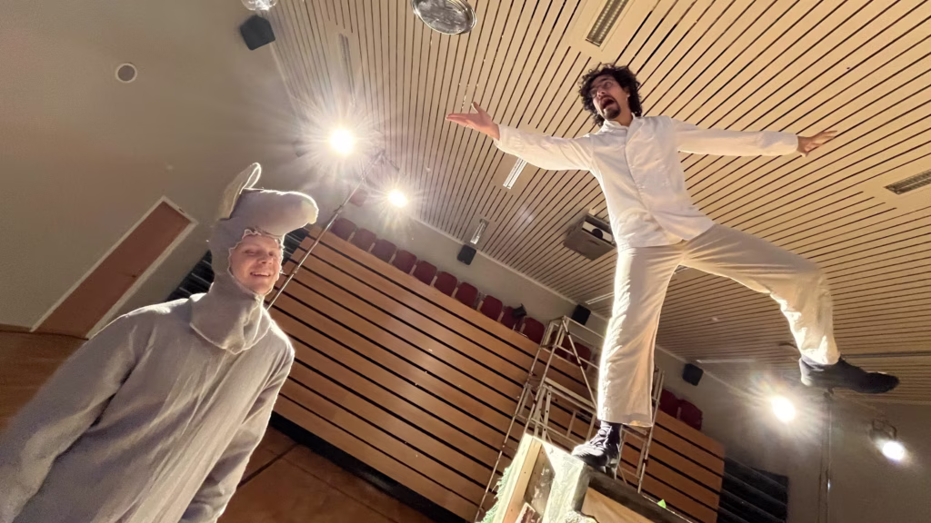 En skuespiller i hvit skjorte og beige bukser står på en trapp oppe til høyre. Nede til venstre er en annen skuespiller i et grått rottekostyme.