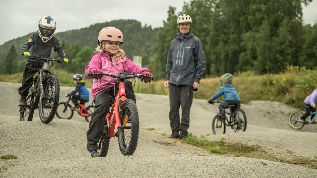 barn i alderen 5-11 år øver på å sykle på en grusbane med større humper og upent terreng. voksen instruktør står ca midt i bildet og passer på.