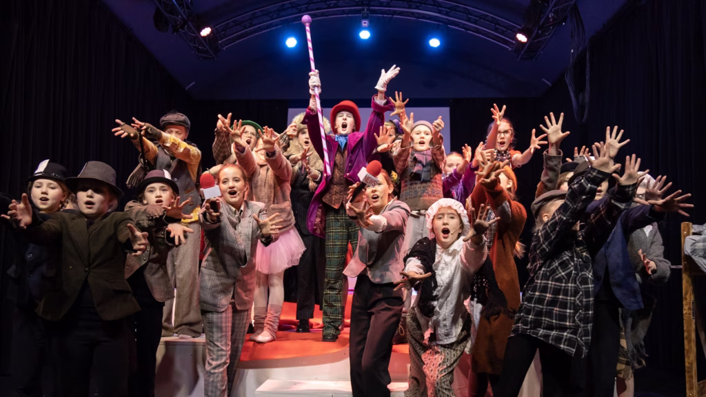 Masse barn som spiller i Charlie og sjokoladefabrikken, som strekker armene mot oss mens de synger. I midten er en kledd ut som Willy Wonka, med lilla frakk, rød hatt og en polkagris-stav. Barna rundt er kledd i dresser, hatter, vester og andre kostymer som minner om andre halvdel av 1800-tallet.